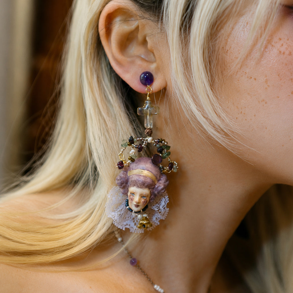 Odette doll-face earring worn beside blonde hair, showing its lilac hair and pale bead drop.