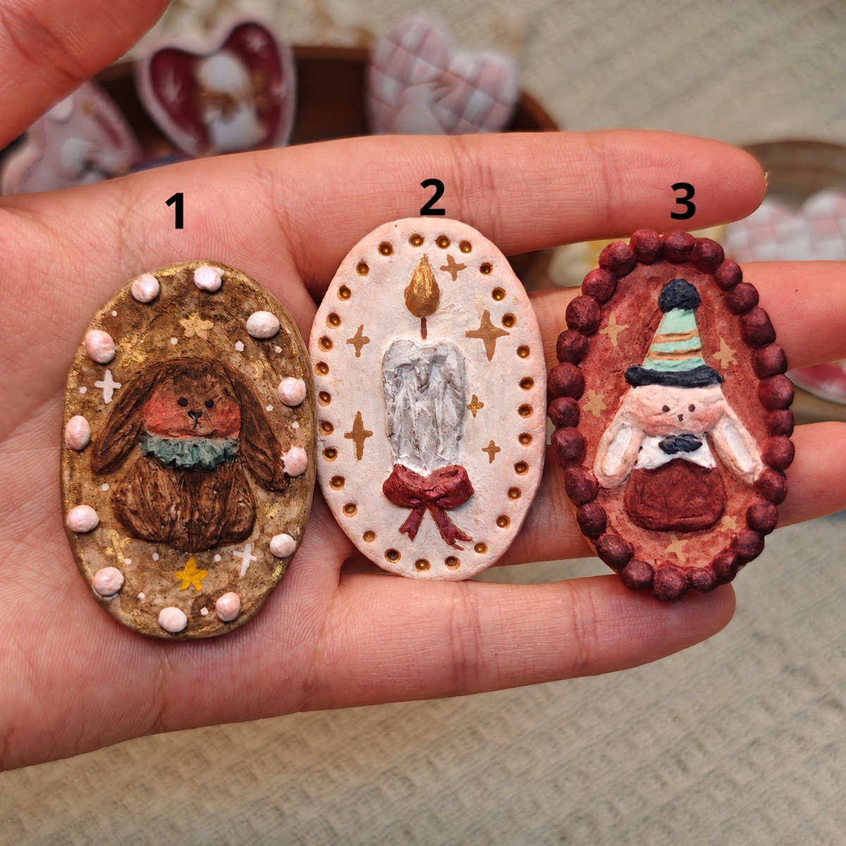Three oval Winterwood Storybook brooches with mushroom, candle and pointed-hat character designs, held in a hand.