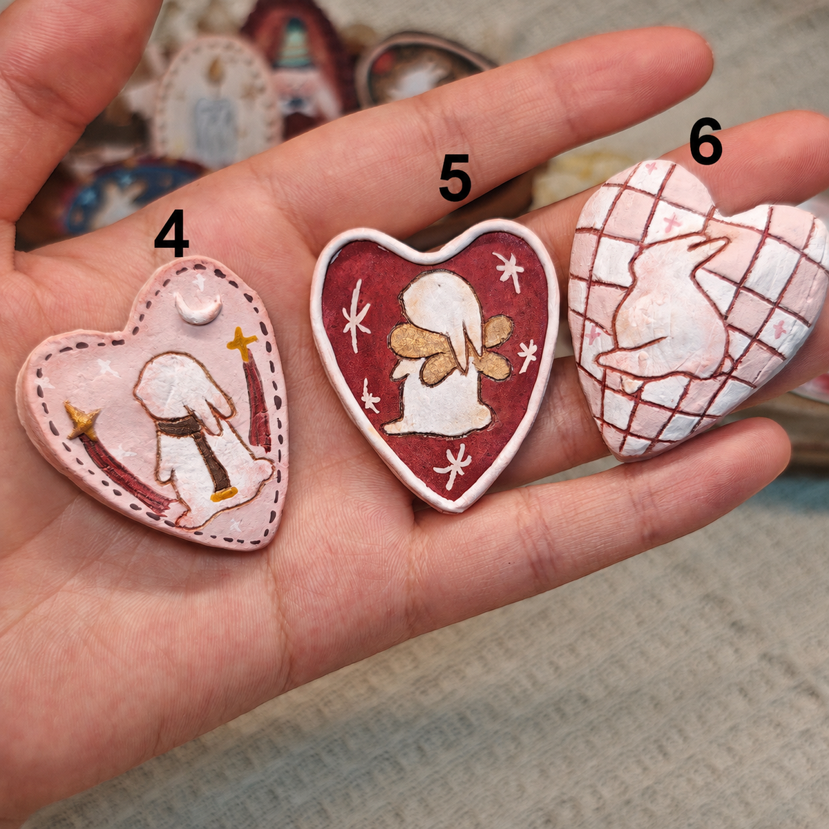 Three heart-shaped Winterwood brooches with pink, red and pale geometric designs, held in a hand.