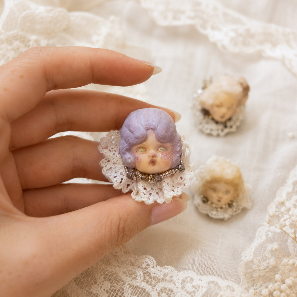 Lambwick doll ring with lilac hair and a cream lace collar, held against white lace.