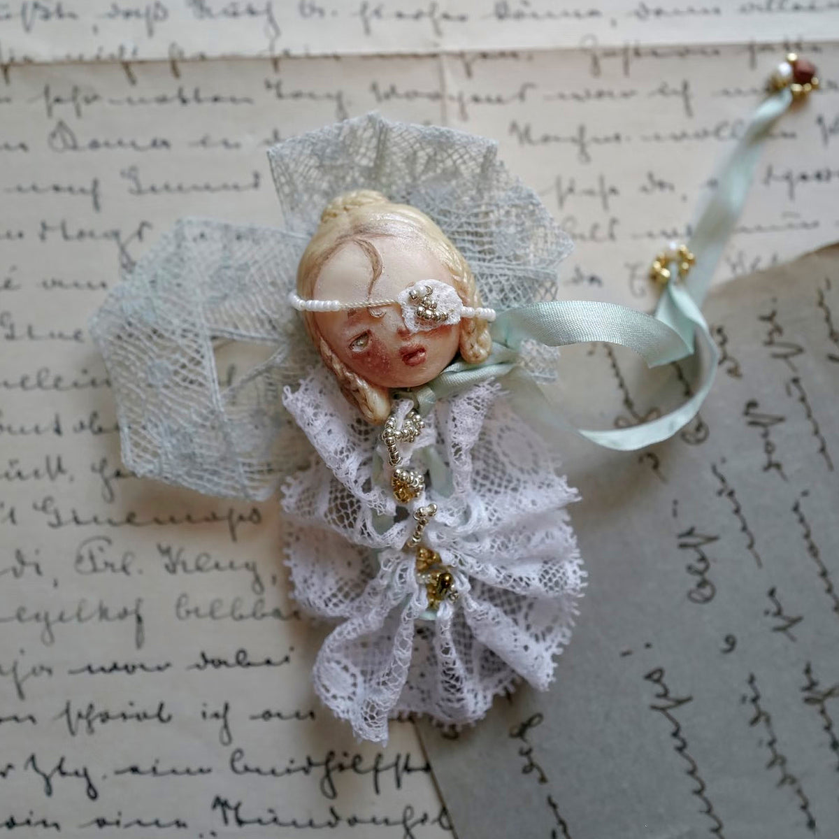 Masked Lambwick doll brooch with white lace, a pale ribbon and a mint-colored bow.