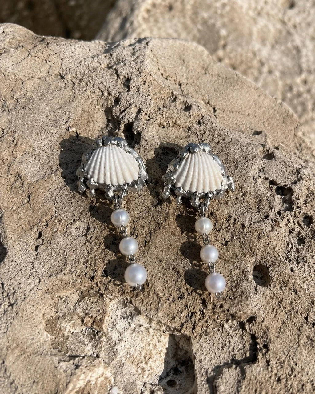 Two Shell Pearl Drop pieces with pale fan shells and hanging beads arranged on a beach rock.