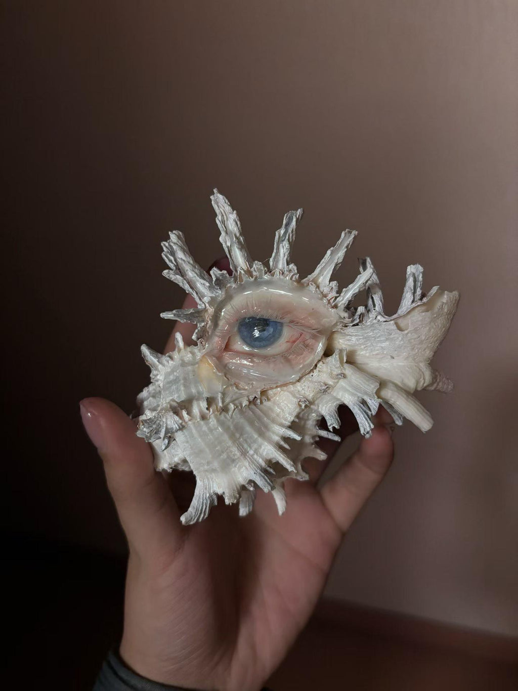 White spiked shell-eye piece held in a hand, showing a pale blue iris and hanging bead drop.