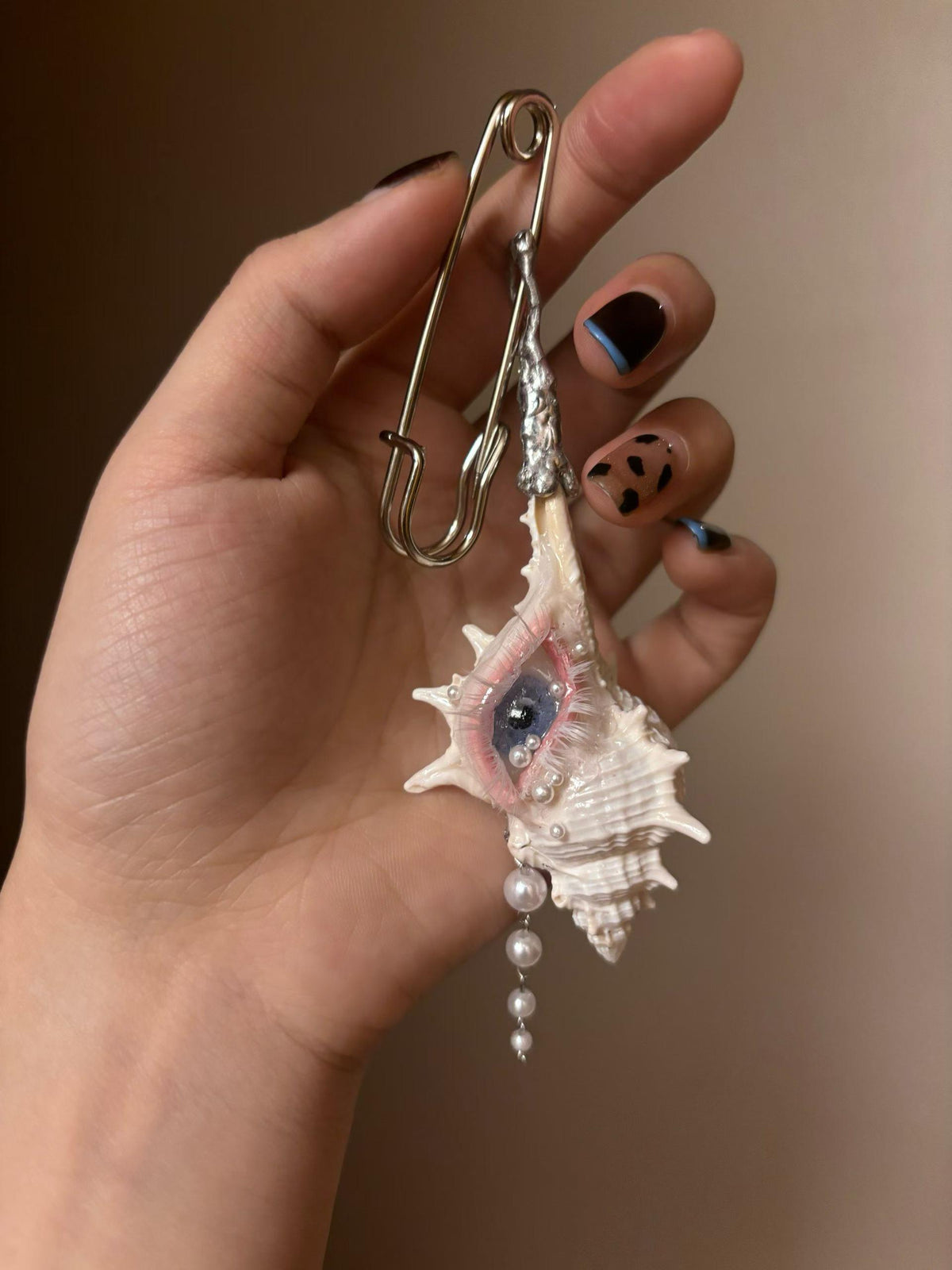 Conch Eye Talisman attached to a large silver-tone safety pin, held in a hand.
