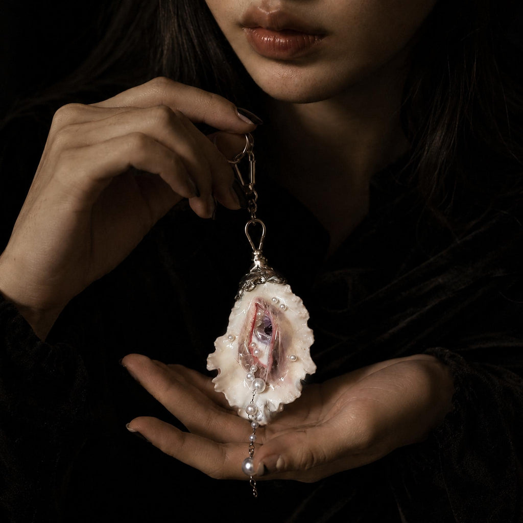 Nyra Ash Grey-Lash oyster eye keychain held between both hands below a model's face.