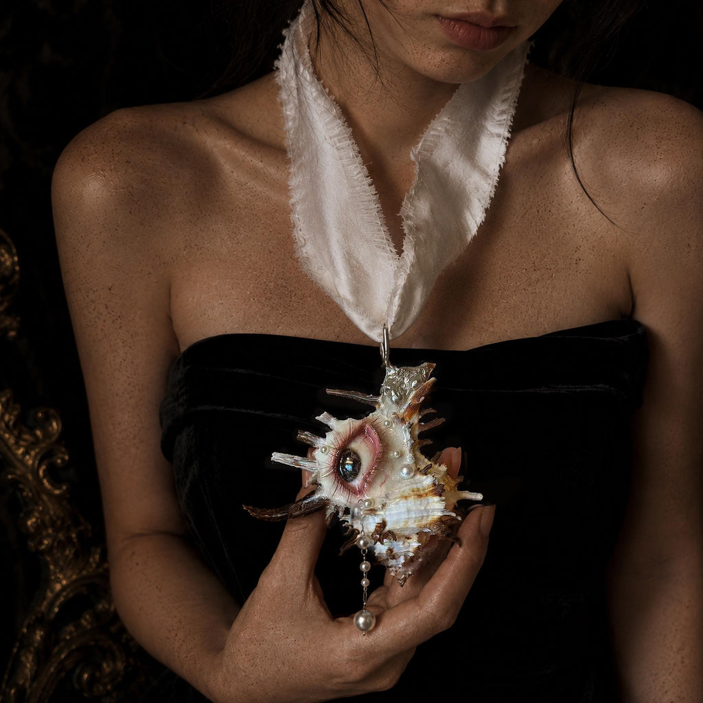 Abyss Guardians shell-eye necklace on ivory ribbon worn over a black dress and held in a hand.