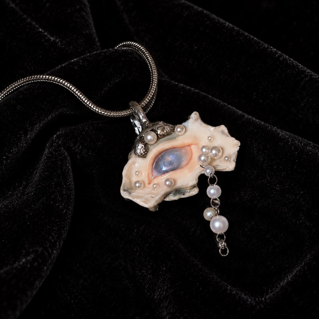 Nyra shell-eye pendant with pale bead drops on a fine metal chain, resting on black velvet.