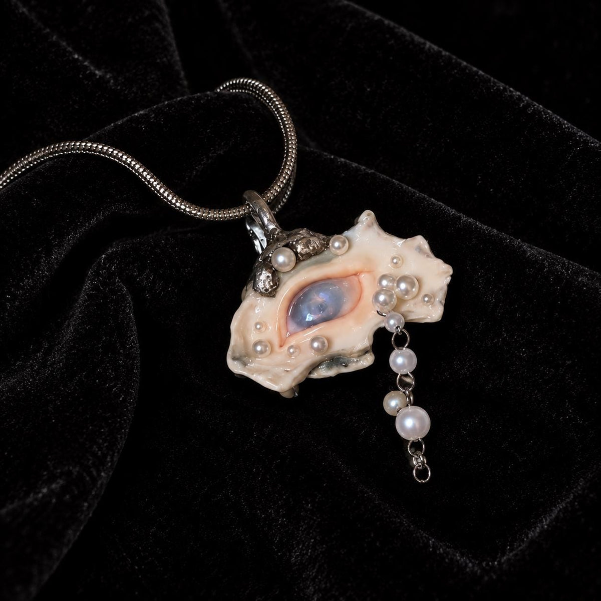 Nyra shell-eye pendant with pale bead drops on a fine metal chain, resting on black velvet.